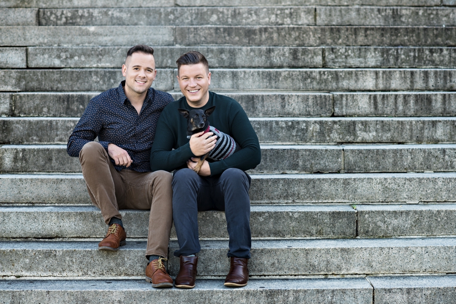 Two grooms to be hold their dog while sitting on the steps in FDR park in Philly for their engagement session.